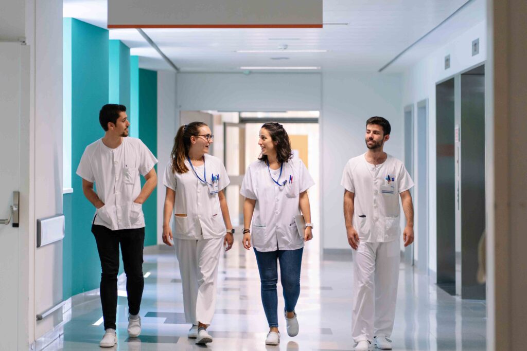 Ein Team aus internationalen Pflegekräften in weißer Arbeitskleidung geht lächelnd durch einen Krankenhausflur.