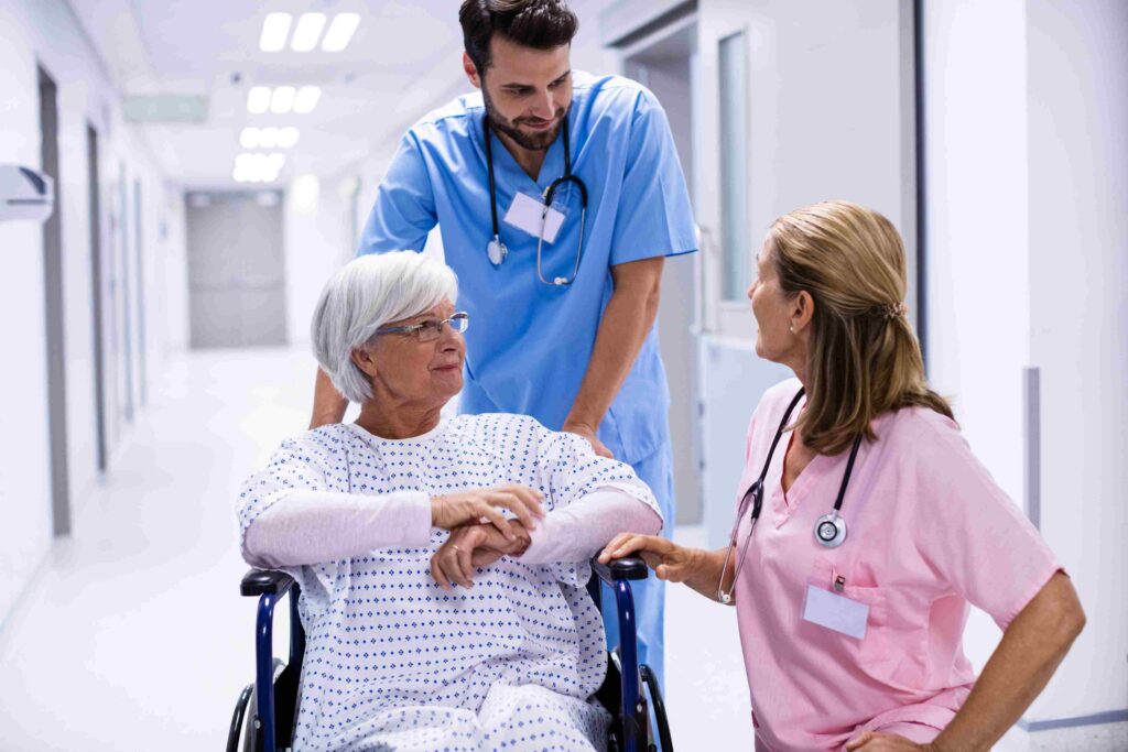 Pflegekräfte sprechen mit einer Seniorin im Rollstuhl im Krankenhausflur.